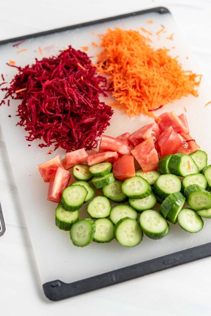 Chopped cucumber, chopped tomato, grated beet and carrot on a cutting board.