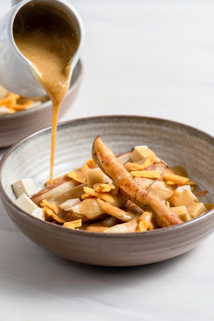 Pouring gravy over baked fries, tofu and cheese in a bowl.