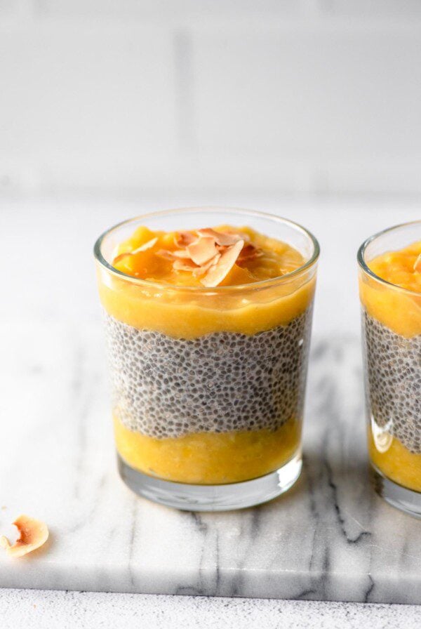 A glass jar of layered mango chia pudding topped with coconut flakes. Another jar of pudding is in the background.