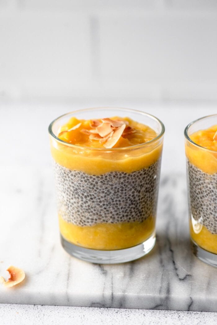 A glass jar of layered mango chia pudding topped with coconut flakes. Another jar of pudding is in the background.