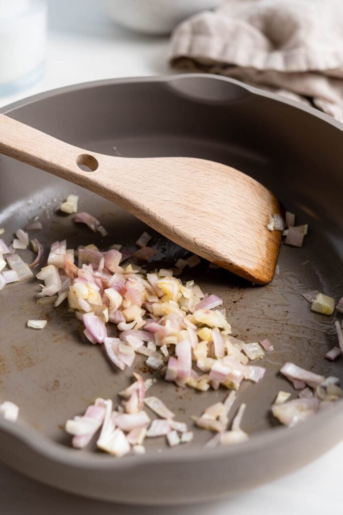 Minced shallots being cooked in a saucepan with a wooden spoon.