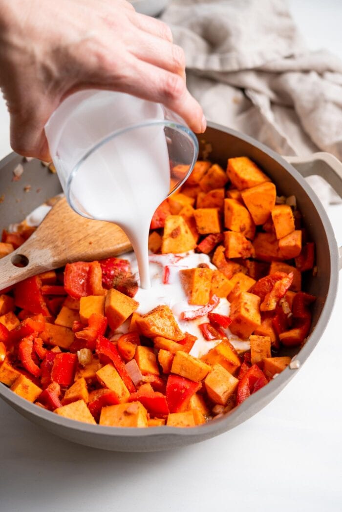 Pouring a jar of coconut milk into a skillet of sweet potato and bell peppers.