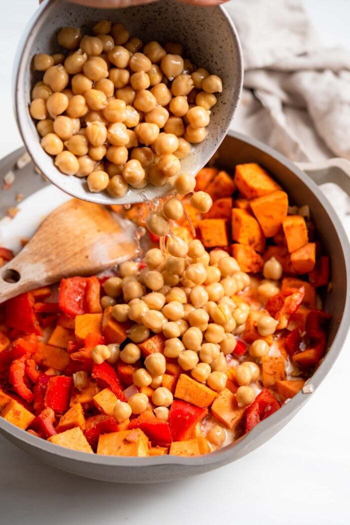 Dumping a bowl of chickpeas into a skillet of sweet potato and bell pepper.