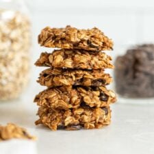 Stack of 5 oatmeal chocolate chips cookies with jars of oats and chocolate chips in the background.