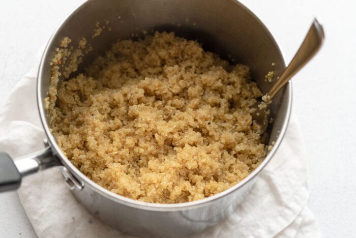 A pot of fluffy cooked quinoa with a fork resting in it. The pot is sitting on a folded dish cloth.