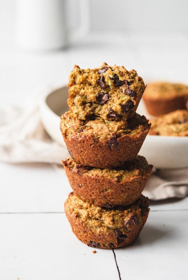 Stack of four chocolate chip zucchini muffins. Muffin on top is cut in half to show inside texture.