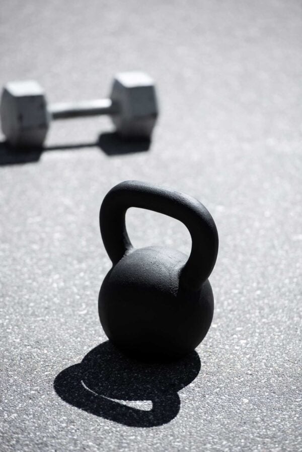 Black kettlebell on pavement with a dumbbell behind it.