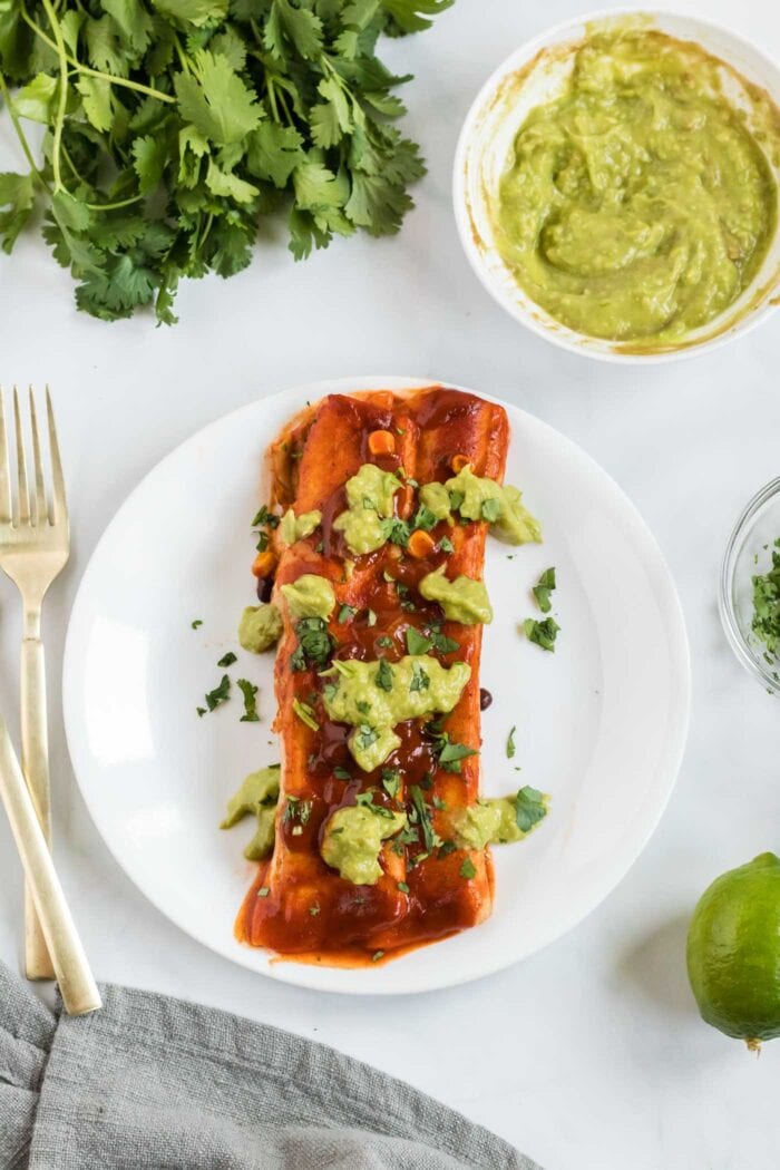 An enchilada topped with avocado sauce on a small plate with a fork and bowl of avocado sauce beside it.