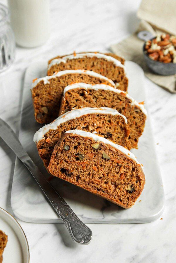 Sliced vegan carrot cake banana bread topped with cream cheese frosting on a marble cutting board.