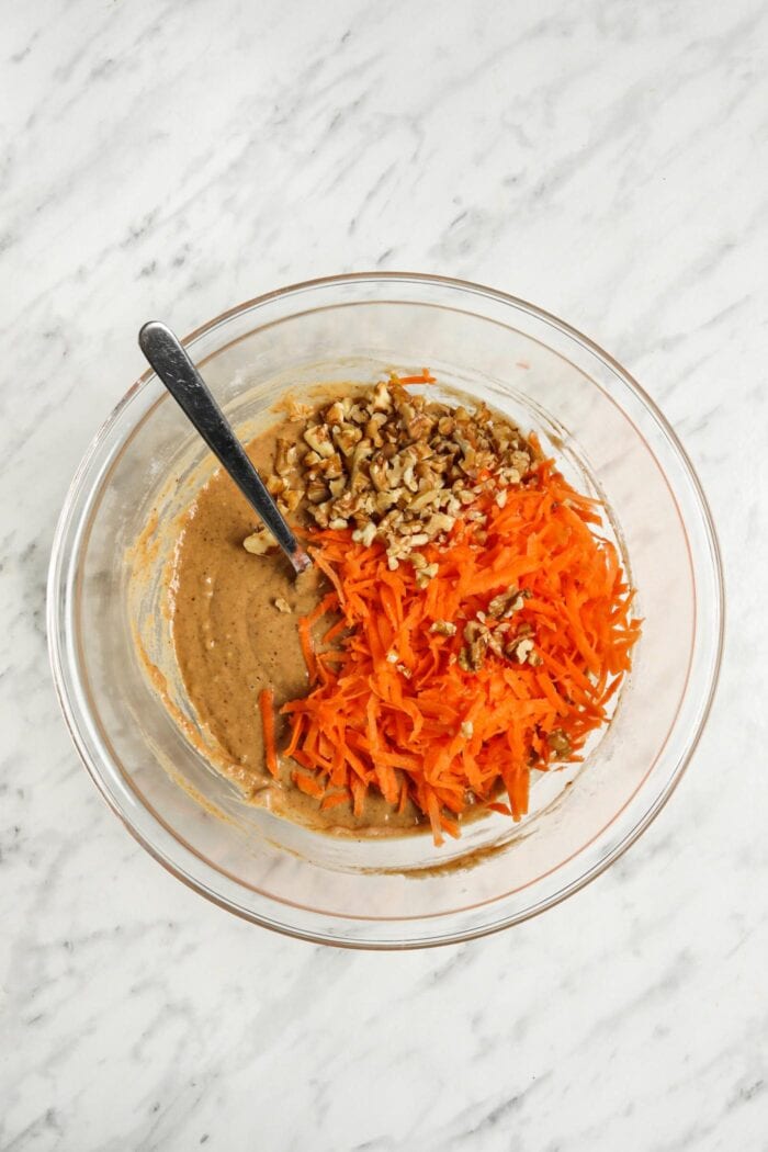 Grated carrot and walnuts in a glass mixing bowl of raw batter.