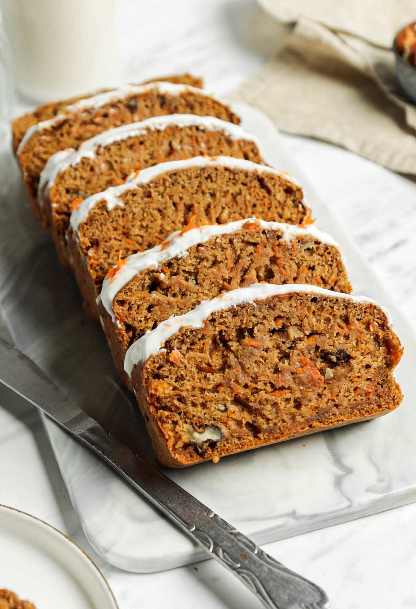 Sliced vegan carrot cake banana bread topped with cream cheese frosting on a marble cutting board.