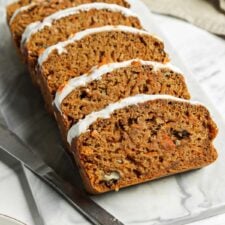 Sliced vegan carrot cake banana bread topped with cream cheese frosting on a marble cutting board.