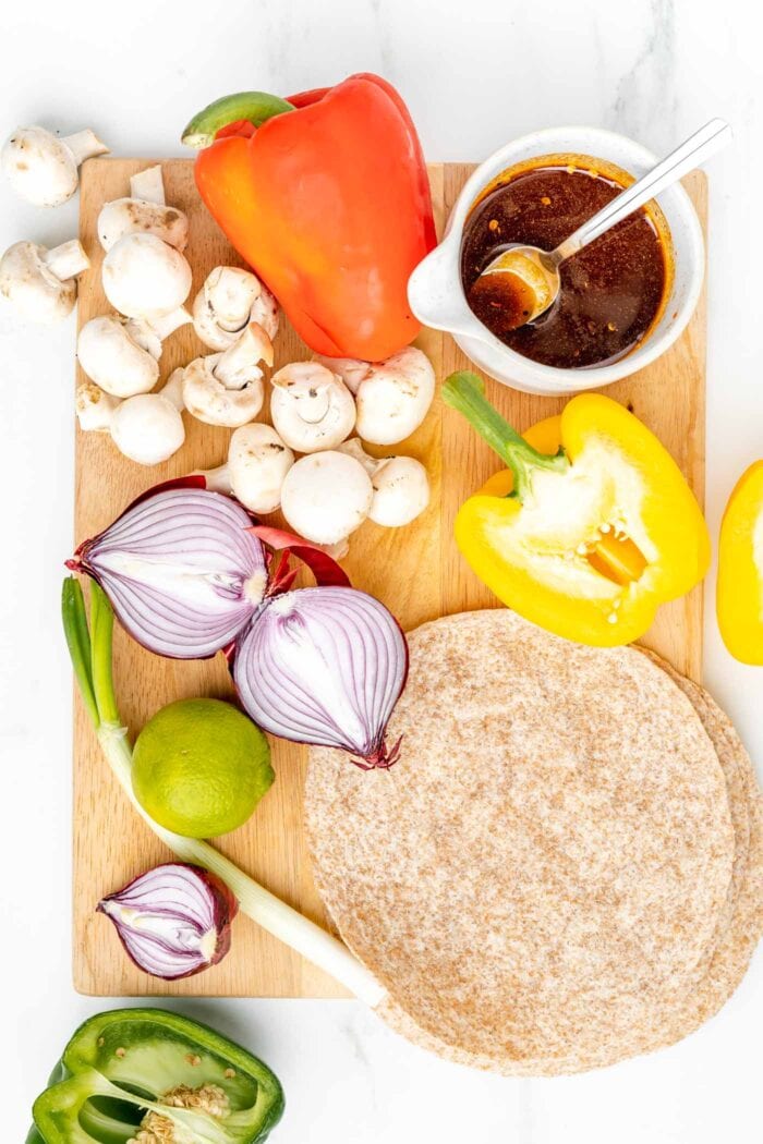 Overhead view of various vegetables on a cutting board with some marinade in a bowl and a wheat tortilla.