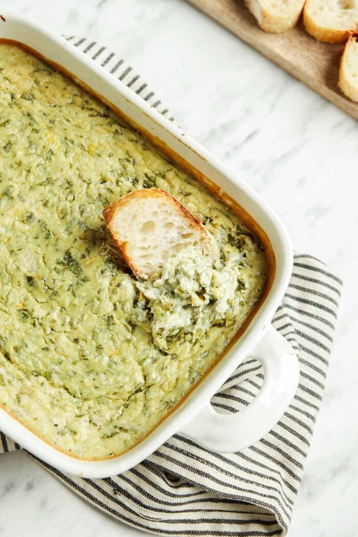 Dipping some bread into a baking dish of spinach artichoke dip.