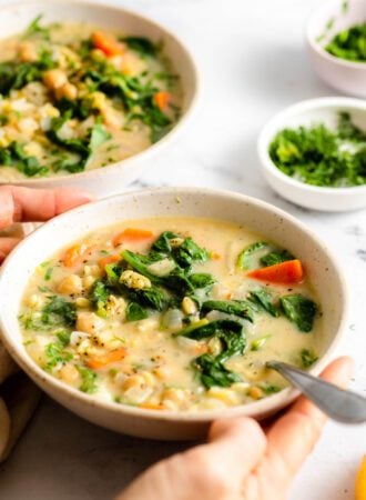 Two hands holding a bowl of Greek lemon chickpea orzo soup with spinach in it. A spoon rests in the bowl.