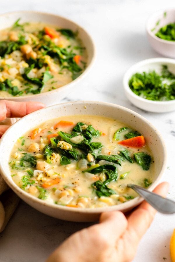 Two hands holding a bowl of Greek lemon chickpea orzo soup with spinach in it. A spoon rests in the bowl.