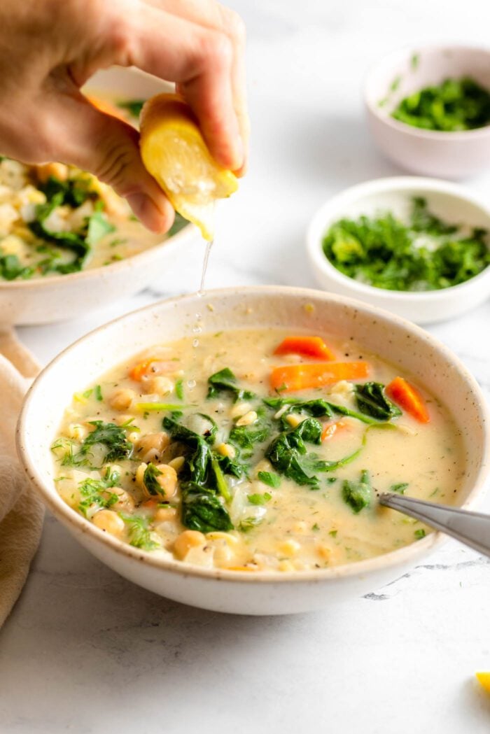 Hand squeezing a lemon wedge over a bowl of lemon chickpea orzo soup with spinach and carrots.
