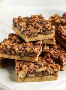 Stack of pecan squares on a small cutting board. Jar of coffee and stack of plates are in the background.