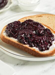 Piece of toast topped with blueberry chia jam. Jar of blueberry jam and a mug of coffee in the background.