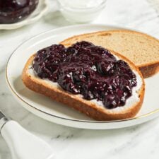 Piece of toast topped with blueberry chia jam. Jar of blueberry jam and a mug of coffee in the background.