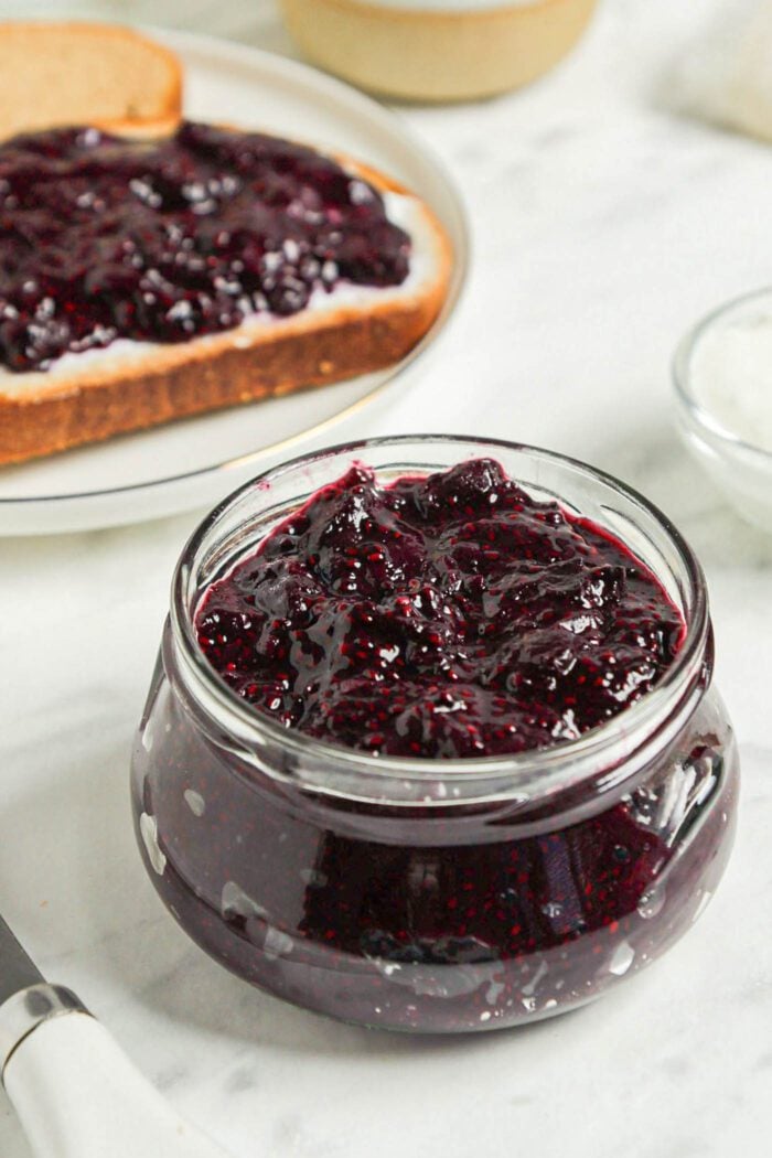 Small jar of blueberry chia seed jam with a piece of bread topped with jam in the background.