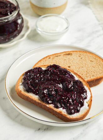 Piece of toast topped with blueberry chia jam. Jar of blueberry jam and a mug of coffee in the background.