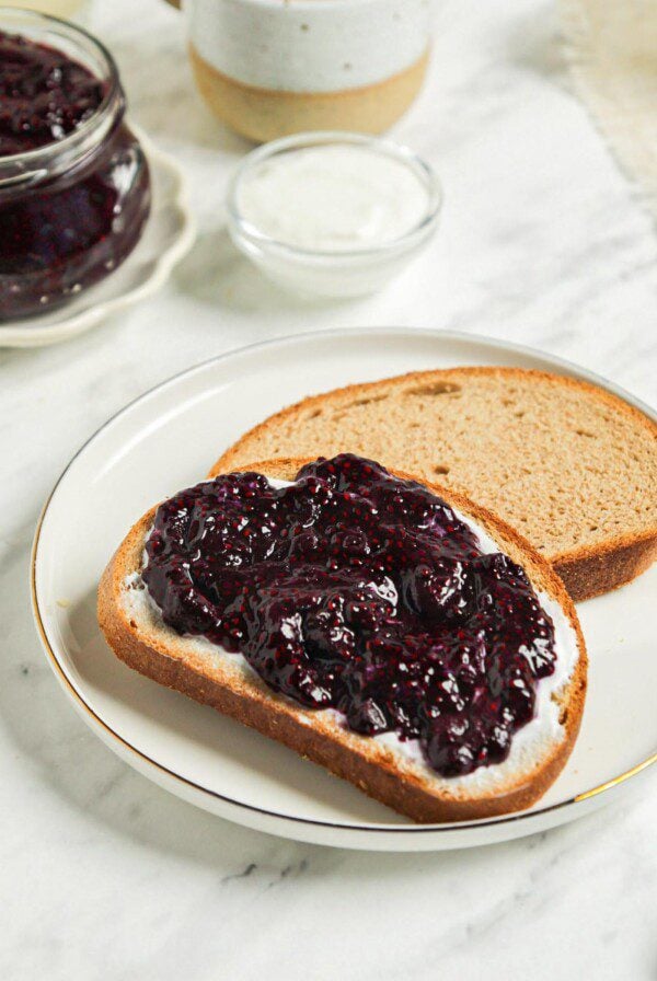 Piece of toast topped with blueberry chia jam. Jar of blueberry jam and a mug of coffee in the background.