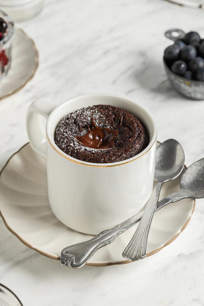 Chocolate mug cake on a small plate with 2 spoons resting on plate.
