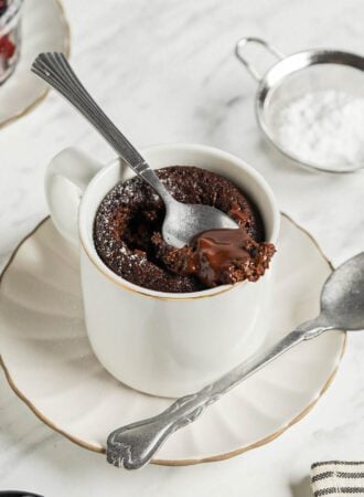 Spoon taking a spoonful of chocolate mug cake from a mug sitting on a small plate.