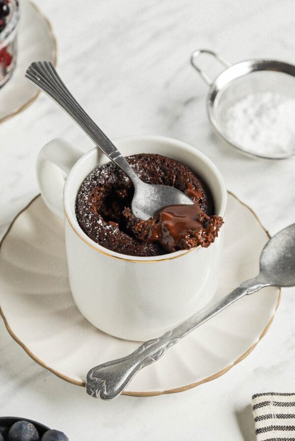 Spoon taking a spoonful of chocolate mug cake from a mug sitting on a small plate.