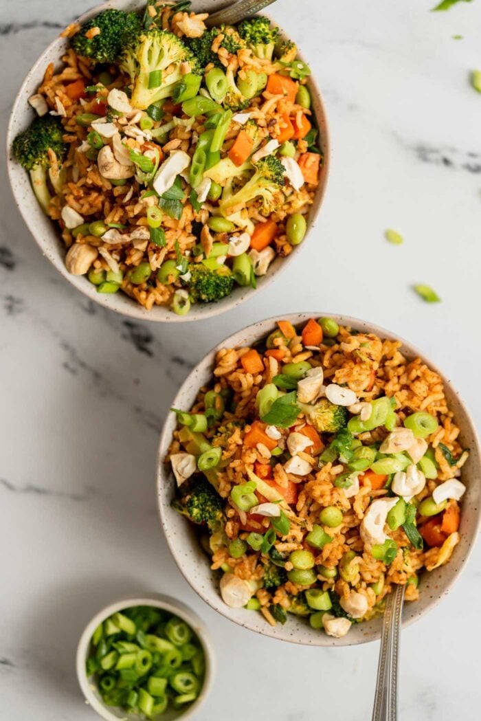 Two bowls of fried rice with broccoli, carrot, edamame and green onion topped with chopped cashews.