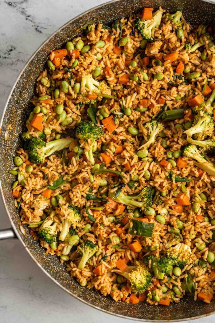 Curry fried rice with vegetables and edamame cooking in a large skillet with a wooden spoon.