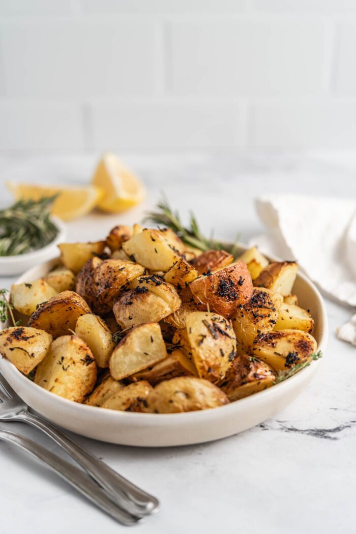 Bowl of crispy roasted lemon rosemary potatoes. Two wedges of lemon and some sprigs of rosemary are in the background.
