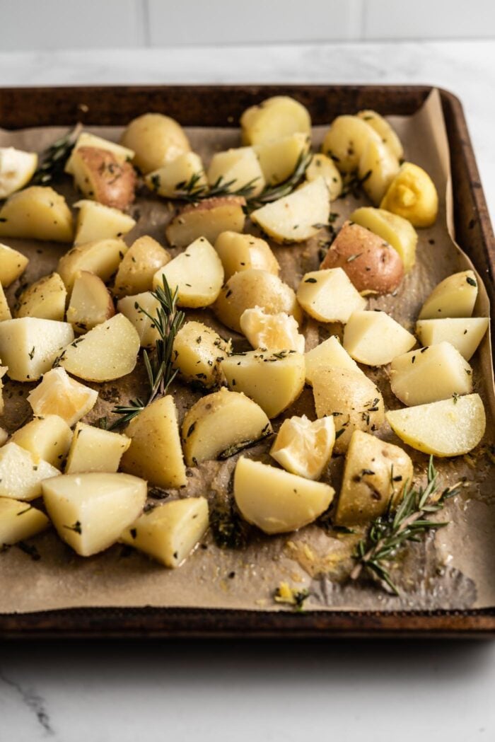 Chopped potatoes on a parchment paper-lined baking tray with spices, lemon and roasemary.