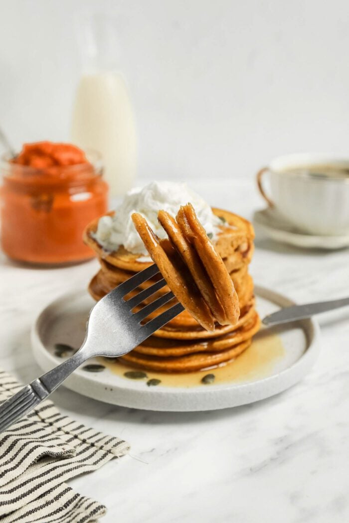 Forkful of pieces of pumpkin pancakes in front of a stack of pumpkin pancakes on a plate.