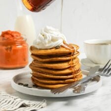 Hand pouring maple syrup from a jar over a stack of pumpkin pancakes topped with whipped cream.