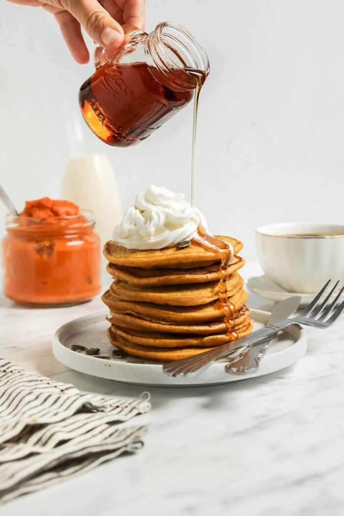 Hand pouring maple syrup from a jar over a stack of pumpkin pancakes topped with whipped cream.