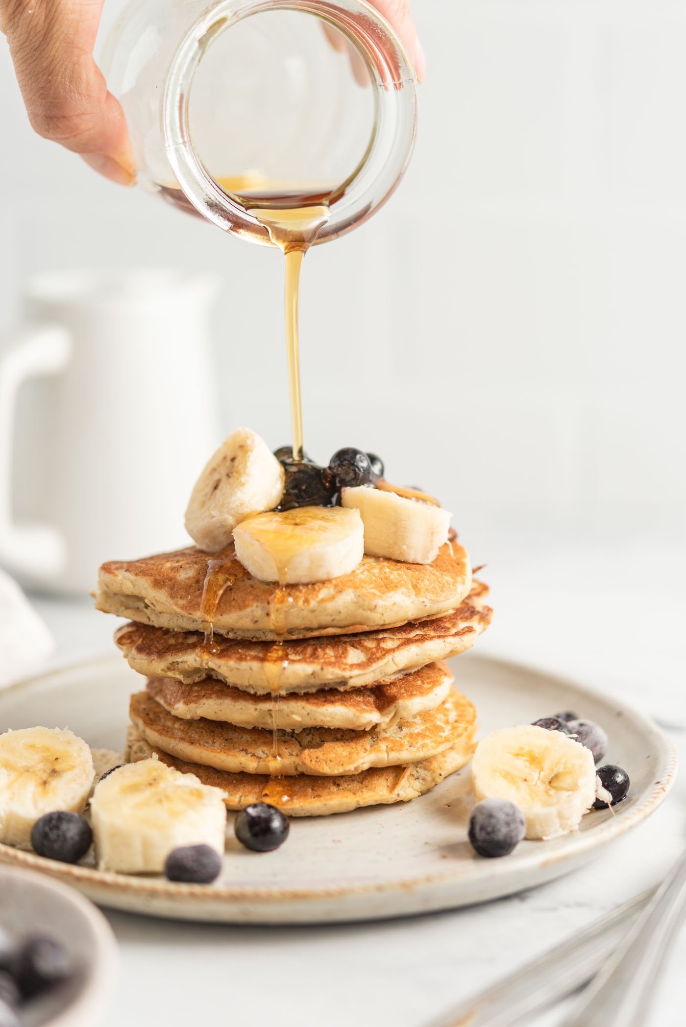 Hand pouring maple syrup from a small jar onto a stack of quinoa flour pancakes topped with banana and blueberry.