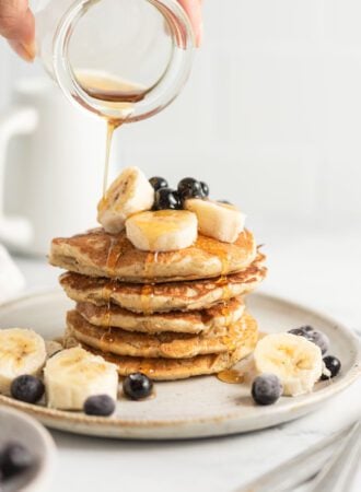 Hand pouring maple syrup from a small jar onto a stack of quinoa flour pancakes topped with banana and blueberry.
