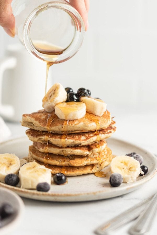 Hand pouring maple syrup from a small jar onto a stack of quinoa flour pancakes topped with banana and blueberry.