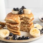 Close up of a stack of pancakes that have been sliced to show the texture inside. The pancakes are topped with blueberries and banana.