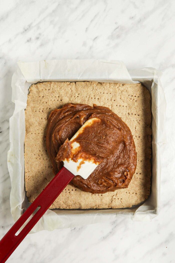 Spatula spread date caramel over a shortbread cookie crust in a pan.