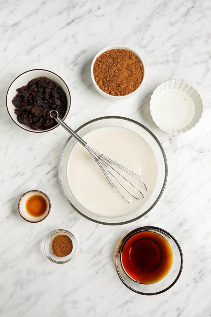 A bowl of milk with a whisk in it beside bowls of maple syrup, raisins and coconut sugar.