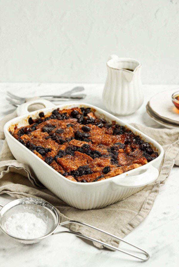 Baked bread pudding in a baking dish. A mesh strainer rests beside dish.