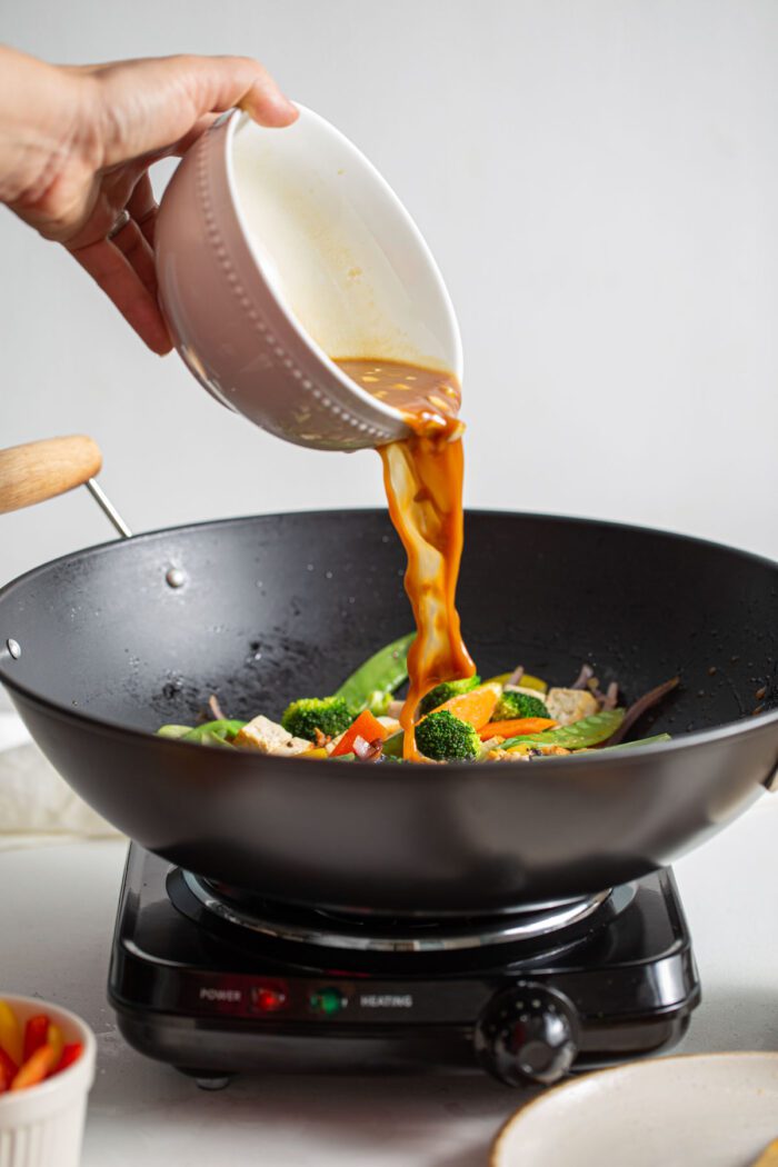 Pouring teriyaki sauce over vegetables cooking in a wok.