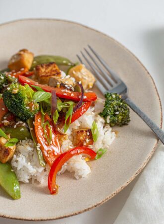 White rice topped with a teriyaki tofu stir fry with bell peppers and broccoli on a small plate with a fork.