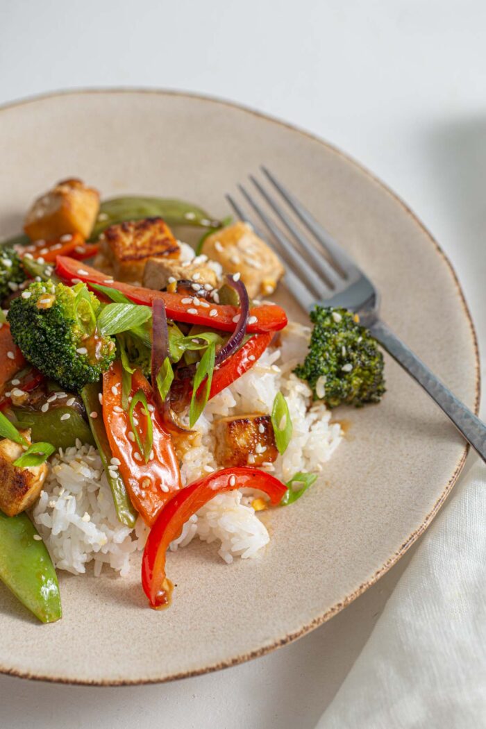 White rice topped with a teriyaki tofu stir fry with bell peppers and broccoli on a small plate with a fork.