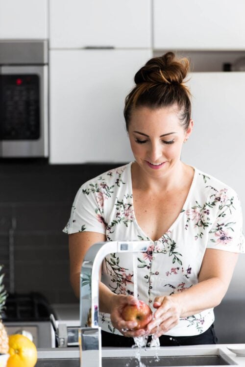 A woman with her hair up wearing a floral shirt washing an apple under a tap.