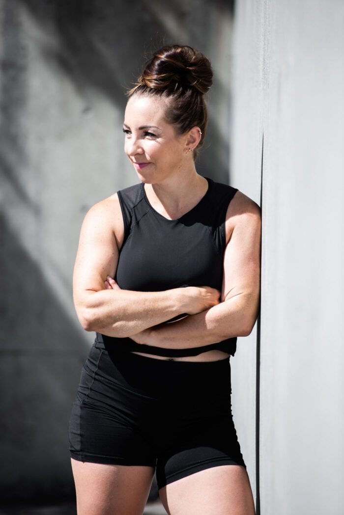 A woman in athletic clothing leaning against a concrete wall looking to the side with her arms crossed.