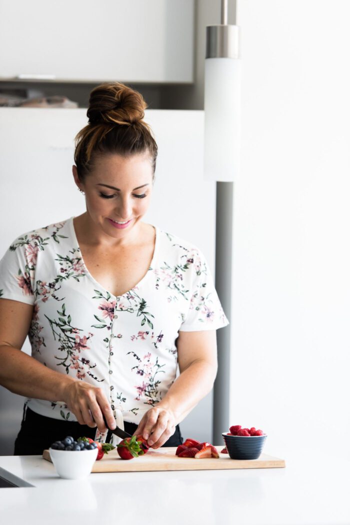 A woman with her hair up wearing a floral shirt cutting strawberries on a cutting board